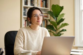Femme portant des lunettes à monture fine travaillant sur son ordinateur portable dans un bureau à domicile chaleureux